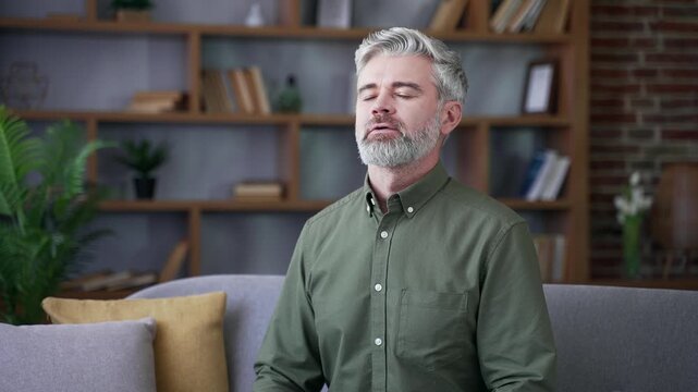 Mature bearded man with gray hair relaxing with eyes closed while sitting on sofa at home. Calm male breathing deeply, feeling peace, happiness, and relief in cozy living room interior after long day