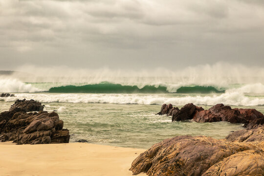 A breaking wave throws up clouds of wind blown sea spray as it thunders towards the rock and sand coastline on a windy day under an overcast sky at Pambula Beach in New South Wales, Australia.