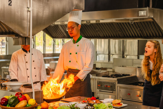 Asian chef cooking with flaming pan as teen culinary interns watch in a professional kitchen