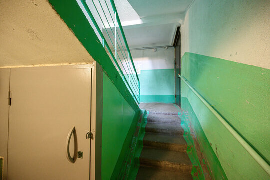 Narrow stairwell features concrete steps, green-painted walls and railings, utilitarian cabinet with handle, worn edges, muted lighting, utilitarian design, corridor layout
