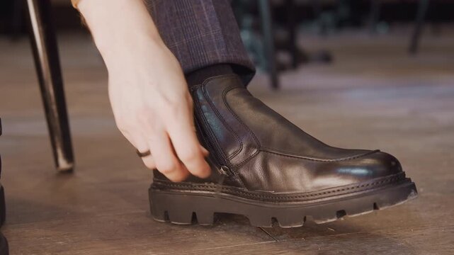 Close up of man foot wearing black leather ankle boot with side zipper as hand adjusts shoe. Boot features smooth texture and thick sole set against wooden floor background with soft