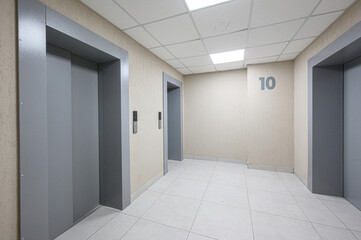 Spacious elevator lobby: beige textured walls, white tiled floor, white ceiling tiles, gray elevator portals, wall-mounted control panels, number 10 on wall, clean functional design © evgeniykleymenov