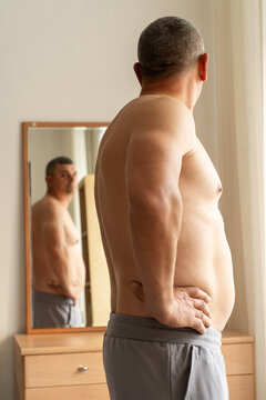 An overweight, middle-aged man stands shirtless with his back to camera, looking at his reflection in mirror and assessing changes in his physique. Real person, self-acceptance, authentic lifestyle