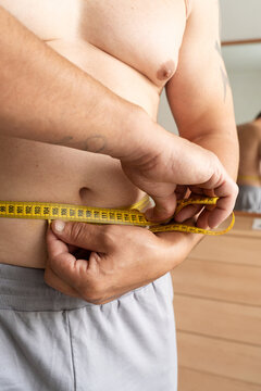 Man measures his waist with tape measure at home, tracking his weight loss progress. Real person, self-acceptance, natural lifestyle, weight control