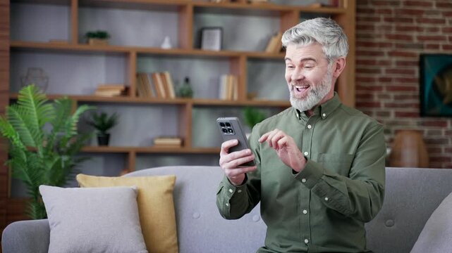 Bearded man with gray hair sitting on sofa at home reading great news on smartphone. Excited male smiling, celebrating positive message and enjoying moment of happiness and relief in cozy living room