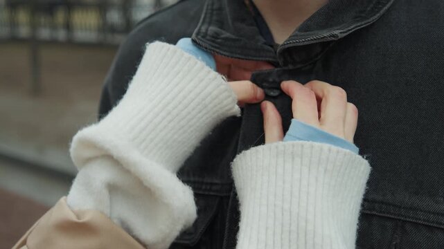 Close up of young woman assisting another woman by buttoning jacket on their dark denim coat. interaction involves hands in white knit sleeves and light blue cuffs focusing on precise manual