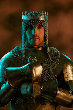 Portrait of young adult Caucasian king wearing armor and crown while holding sword for historical leadership concept, suitable for game marketing, vertical