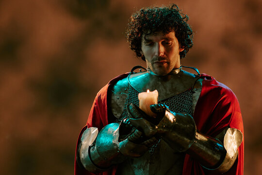 Young adult Caucasian man wearing knight armor holding burning candle thoughtfully. Historical character concept suitable for fantasy game marketing and book cover design
