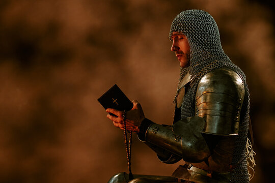 Young adult Caucasian male knight wearing armor holding holy bible and rosary beads while praying. Image suitable for historical marketing, medieval themes, cinema production