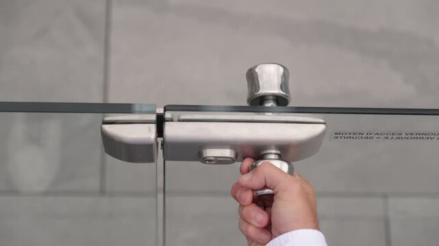 A close-up shows a hand operating the metal latch of a glass pool gate at a holiday club, with the transparent panels against a blurred gray background.