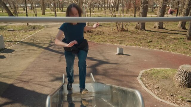 Close up of woman enjoying herself on playground equipment in outdoor park setting. Woman sits and slides down then stands at top preparing to slide again with sunlight casting shadows
