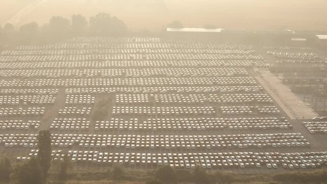 Car storage facility holds thousands of vehicles in dense organized rows across a large industrial yard, with factory structures and morning haze in Italy, drone pullback