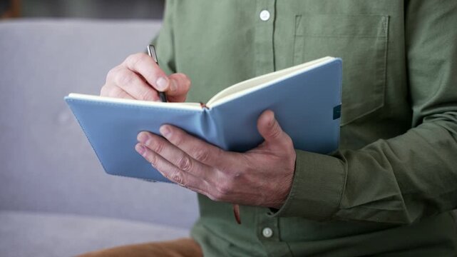 Close-up of male hand writing in notebook while watching online training at home office. Person taking study notes during remote education or webinar, focused on learning and personal development