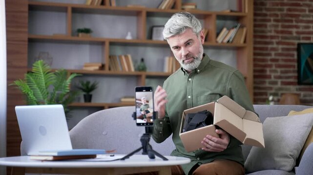 Man with gray hair sitting at home office recording video blog on smartphone while unboxing new product. Influencer live streaming review, sharing opinion, feedback, and advice with online audience