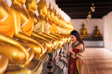 Graceful Asian Woman In Traditional Thai Silk Costume Performing Respectful Wai Prayer Beside Golden Buddha Statues In An Ancient Temple Cloister In Thailand © SHUTTER DIN