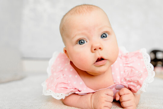 Baby girl 5-6 months old in pink clothes lies on tummy at home and looks at camera, close portrait with blue eyes