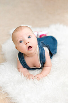 Baby 5-6 months old in blue overalls lies on tummy on a white blanket at home, top view of a development exercise