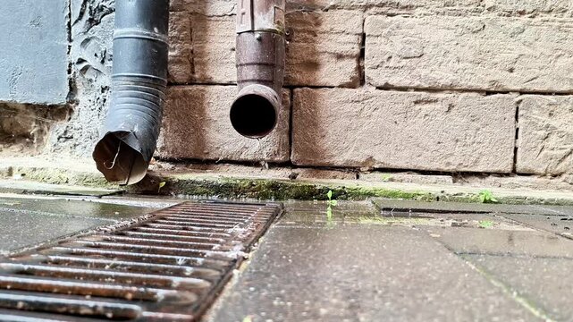 Rainwater flows from drainage pipes into a street grate beside an old stone wall, showing urban stormwater drainage and surface water runoff