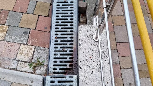 Metal drainage channel with grated cover beside colorful paving stones, concrete sidewalk, and utility pipes, showing urban infrastructure, stormwater management, and pedestrian walkway
