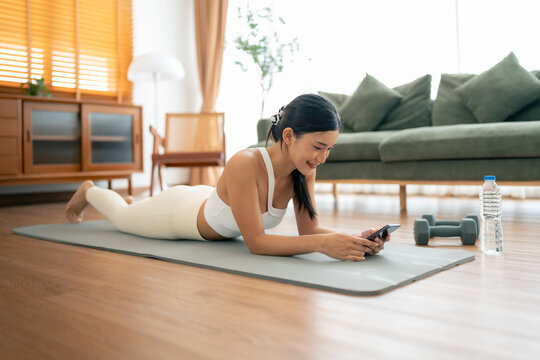Young Asian woman lying on floor on yoga mat using smartphone at home. Beautiful smiling woman doing exercise relaxing while chatting with trainer online after workout