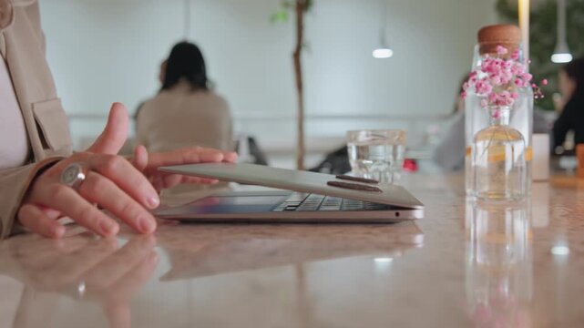 Woman in beige jacket works on laptop at marble table with glass of water and vase flowers other people visible in background. Laptop screen is open as woman interacts by touching keyboard