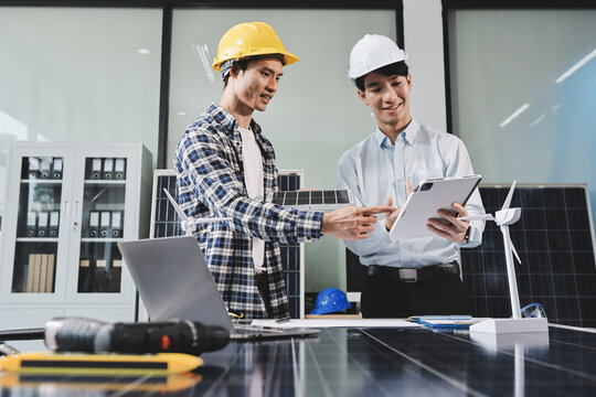 Two Asian engineers wearing hardhats and safety vests discussing solar panel installation and renewable energy project in the office.