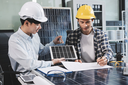 Two Asian engineers wearing hardhats and safety vests discussing solar panel installation and renewable energy project in the office.