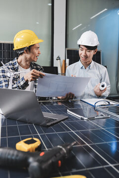 Two Asian engineers wearing hardhats and safety vests discussing solar panel installation and renewable energy project in the office.