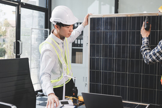 Two Asian engineers wearing hardhats and safety vests discussing solar panel installation and renewable energy project in the office.