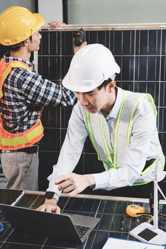 Two Asian engineers wearing hardhats and safety vests discussing solar panel installation and renewable energy project in the office.