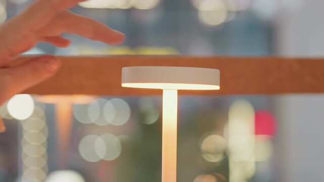 Close up of white touch sensitive lamp with soft light as hand approaches and touches top to turn it on. Lamp remains illuminated when no longer touched set against blurred background