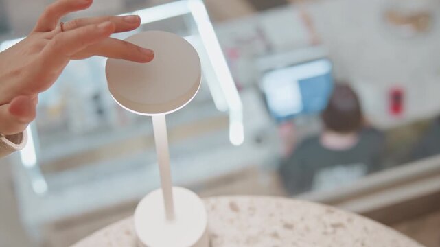 Close up of hand interacting with touch sensitive top surface of elegant white table lamp demonstrating its functionality. background features soft lighting and blurred elements suggesting