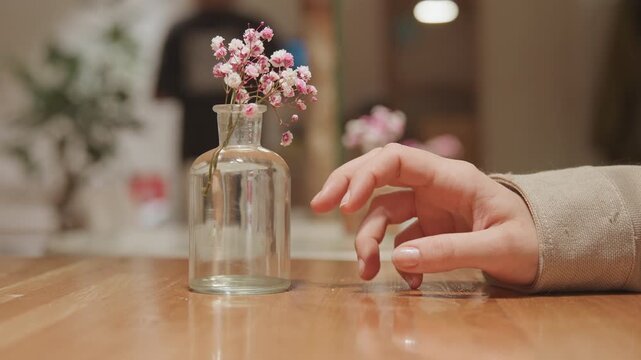 Close up of hand resting on wooden table near clear glass vase with pink flowers soft lighting and blurred background. Hand moves slightly while plate is placed beside vase maintaining focus