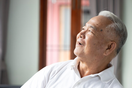 happy elderly southeast asian man in his 70s staying indoors, looking up with hopeful smile in natural window light