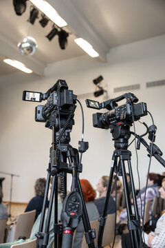 Two professional video cameras on tripods film an indoor event, with attendees softly blurred in the background and camera screens visible