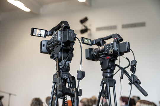 Two professional video cameras on tripods film an indoor event, with attendees softly blurred in the background and camera screens visible