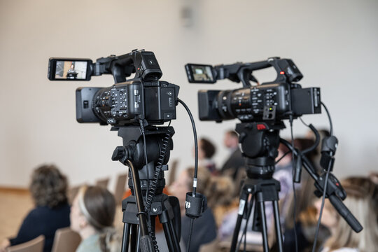 Two professional video cameras on tripods film an indoor event, with attendees softly blurred in the background and camera screens visible