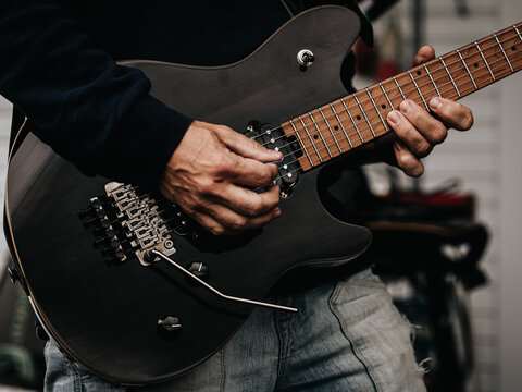 Close-up of a musician playing a black electric guitar, with hands on the strings and fretboard in an indoor setting.