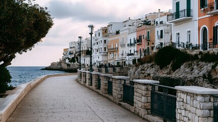Obraz premium A coastal walkway lined with colorful buildings overlooking the sea under a cloudy sky with stone railings and olive trees