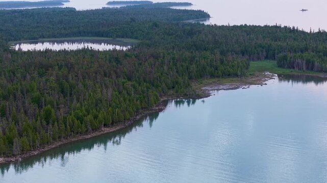 A drone glides beside a densely forested Upper Peninsula shoreline surrounding calm blue water near Hessel Michigan.