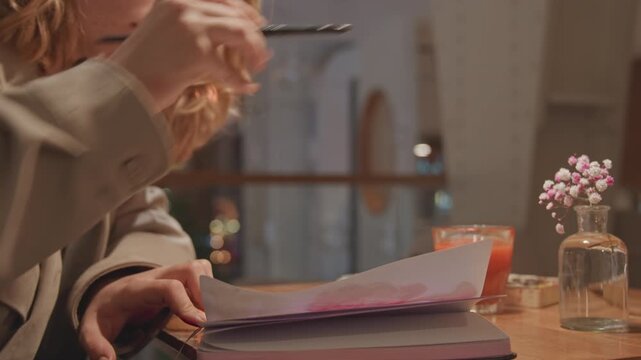Close up of woman painting with watercolors in art journal at wooden table. Woman uses soft brush strokes to create colorful designs including hearts and abstract patterns on white paper