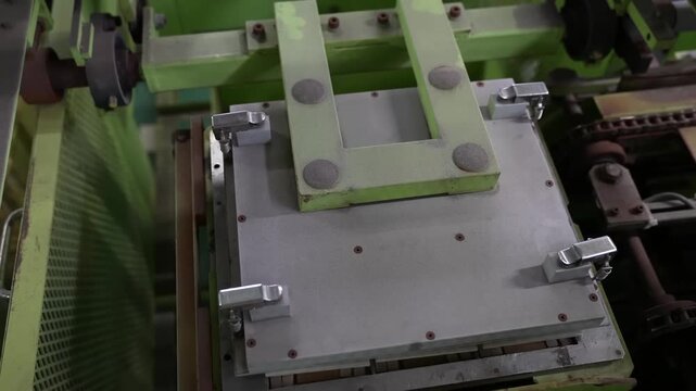 Steel pressing plate lowers onto a tray of pale ceramic tiles inside a green factory machine