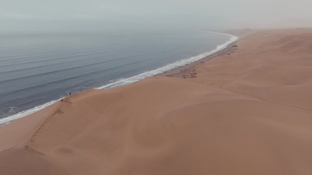 A stunning cinematic aerial shot showing the massive sand dunes of the Namib Desert meeting the crashing waves of the Atlantic Ocean at Sandwich Harbour, Namibia. Ideal for travel and sustainability c