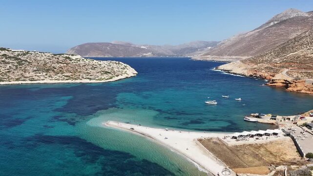 Aerial drone shot flying across the gravel spit of Agios Pavlos beach over clear turquoise reef water with moored boats off Amorgos, Cyclades, Greece, on a sunny day