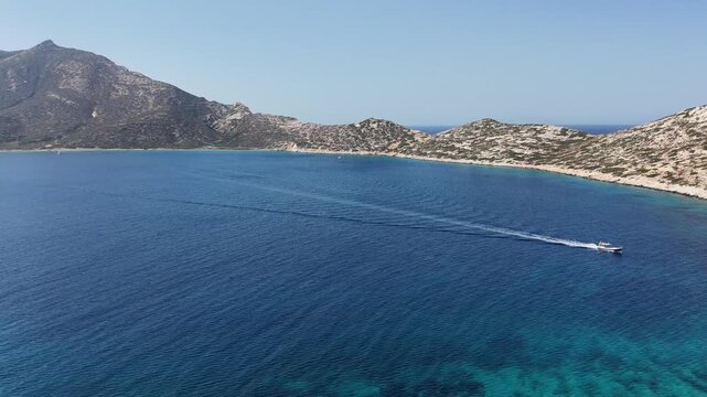 Aerial drone shot turning toward a speedboat leaving a wake trail on the deep blue Aegean Sea below the mountainous islet of Nikouria off Amorgos, Cyclades, Greece, on a sunny day