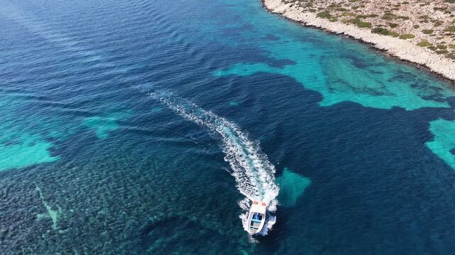 Static aerial drone shot of a speedboat crossing clear turquoise water over a reef near a rocky shore off Amorgos, Cyclades, Greece, on a sunny day