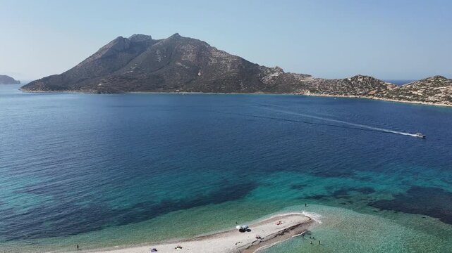 Aerial drone shot of the gravel spit of Agios Pavlos beach reaching into the turquoise Aegean Sea toward the mountainous uninhabited islet of Nikouria off Amorgos, Cyclades, Greece