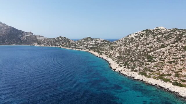 Aerial drone shot tilting up from the blue sea to reveal the arid rocky coastline of the uninhabited islet of Nikouria off the north coast of Amorgos, Cyclades, Greece, on a sunny day