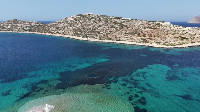 Aerial drone shot flying along the gravel spit of Agios Pavlos beach toward the uninhabited islet of Nikouria across the turquoise Aegean Sea on Amorgos, Cyclades, Greece