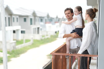 Fototapeta premium Young parents holding their child enjoy the view of the growing suburb from their balcony on the balcony of their new house overlooking the residential estate, shallow depth of field
